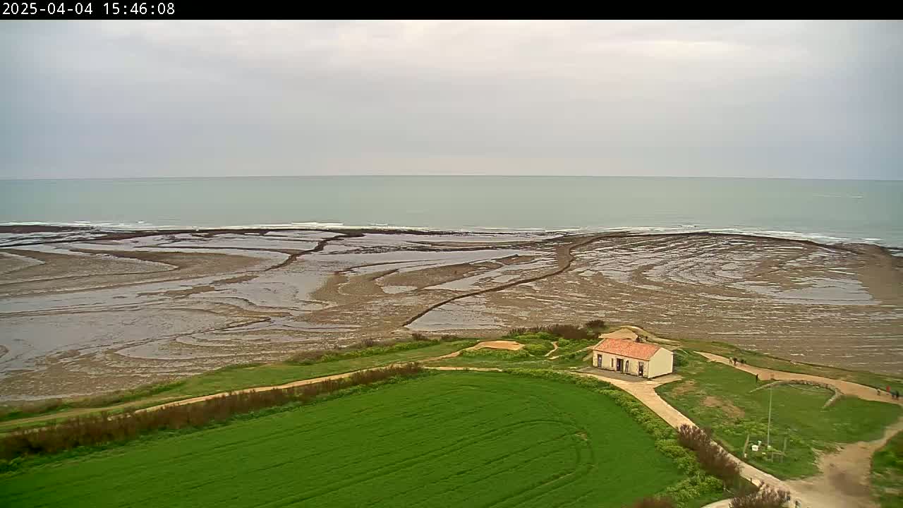 An aerial view shows a small building near a green field overlooking a rocky, tide-exposed coastline and a calm, light-grey sea under an overcast sky.