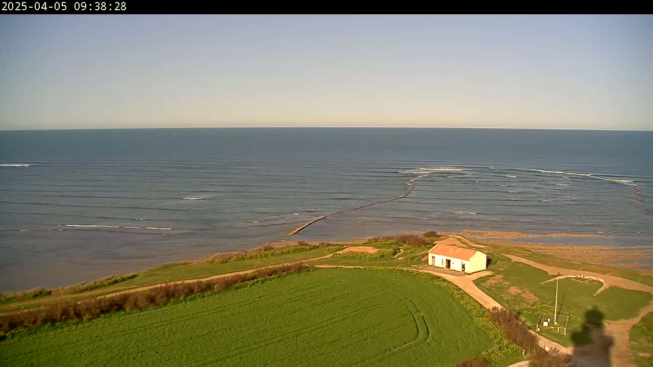 A small building sits on a grassy area overlooking a calm ocean with gentle waves under a clear sky.