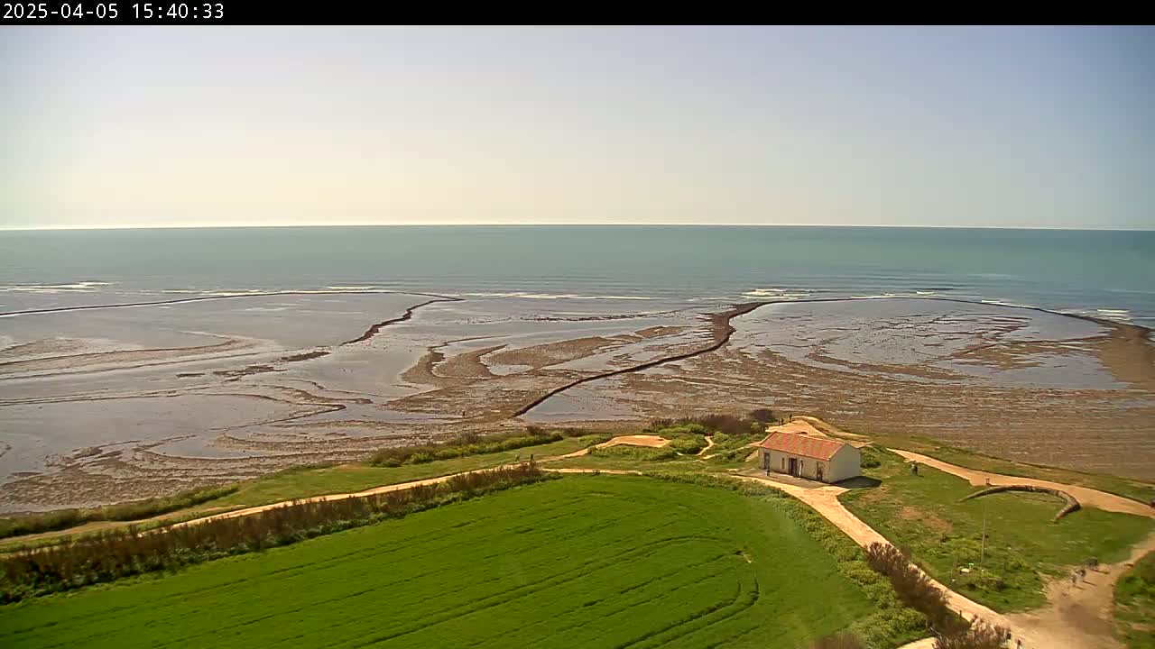 A small, white building sits on a grassy hill overlooking a calm ocean and exposed tidal flats under a mostly clear sky.