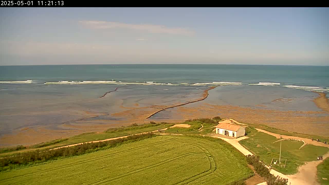 A small, light-colored building sits on a grassy coastal area overlooking a calm ocean with small waves under a partly cloudy sky.