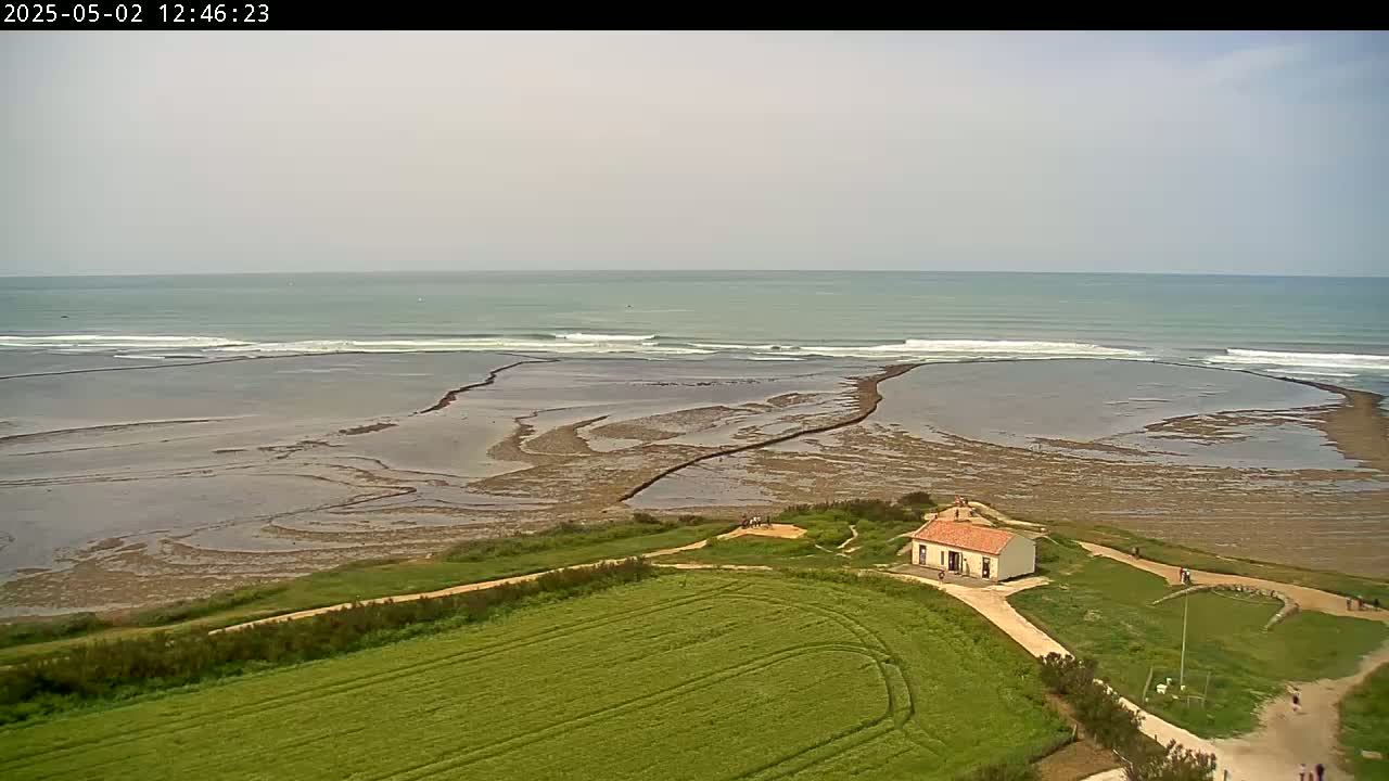 An aerial view shows a small, light-colored building on a grassy area next to a rocky, tidal beach and ocean with small waves under a cloudy sky.