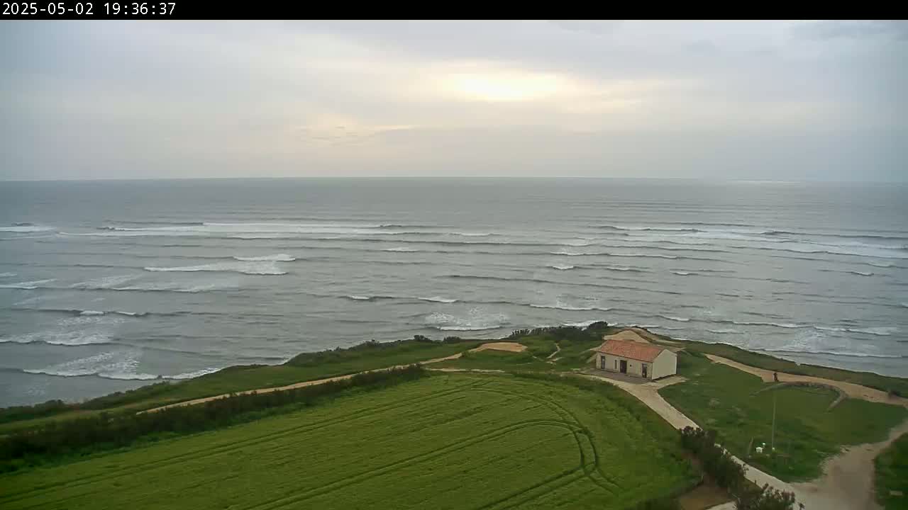 An overcast sky overlooks a small building situated on a grassy coastal area above a gently waving ocean.