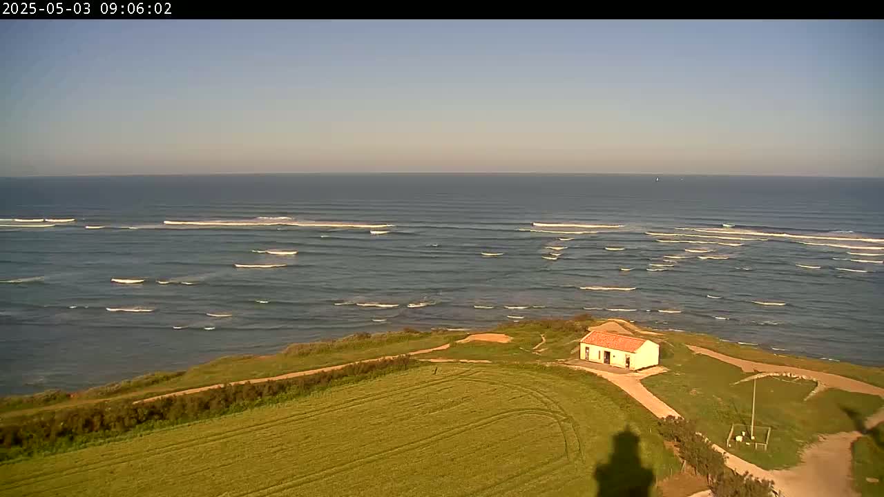 A small, light-colored building with a red roof sits atop a grassy cliff overlooking a calm ocean with small, breaking waves under a clear, light-blue sky.