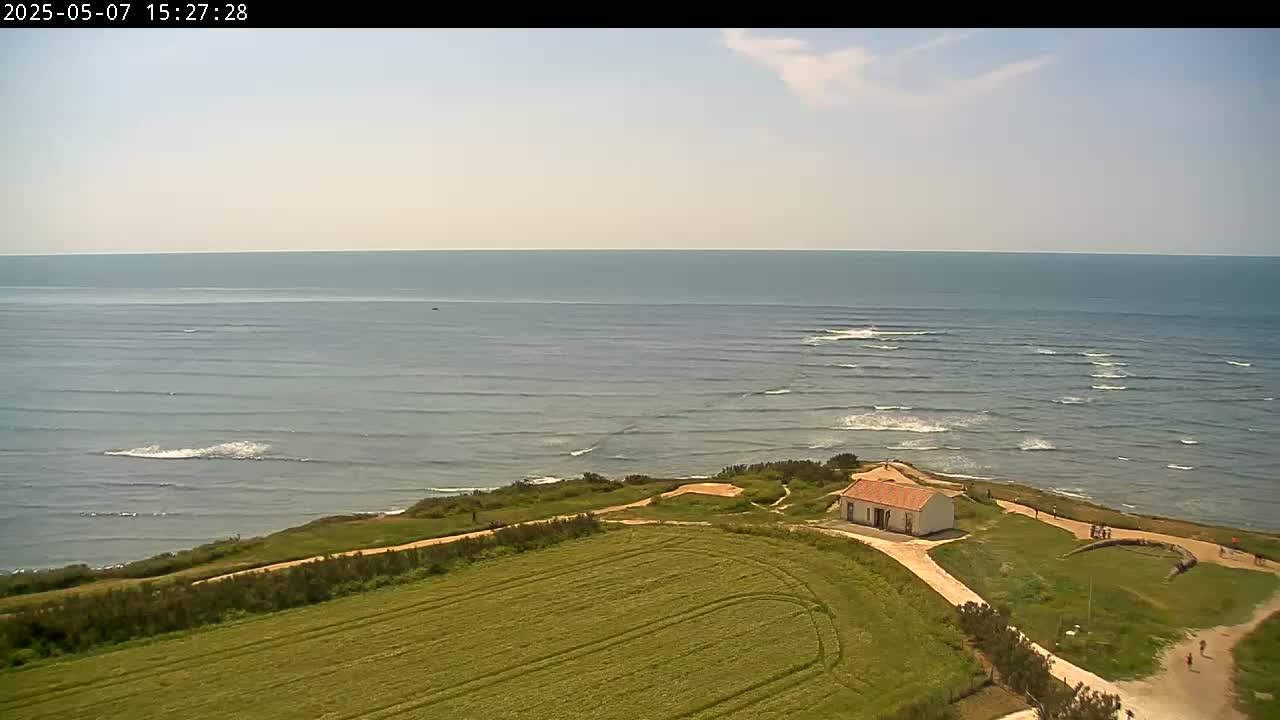 A small, white building sits atop a grassy cliff overlooking a calm ocean with small waves under a mostly clear sky.