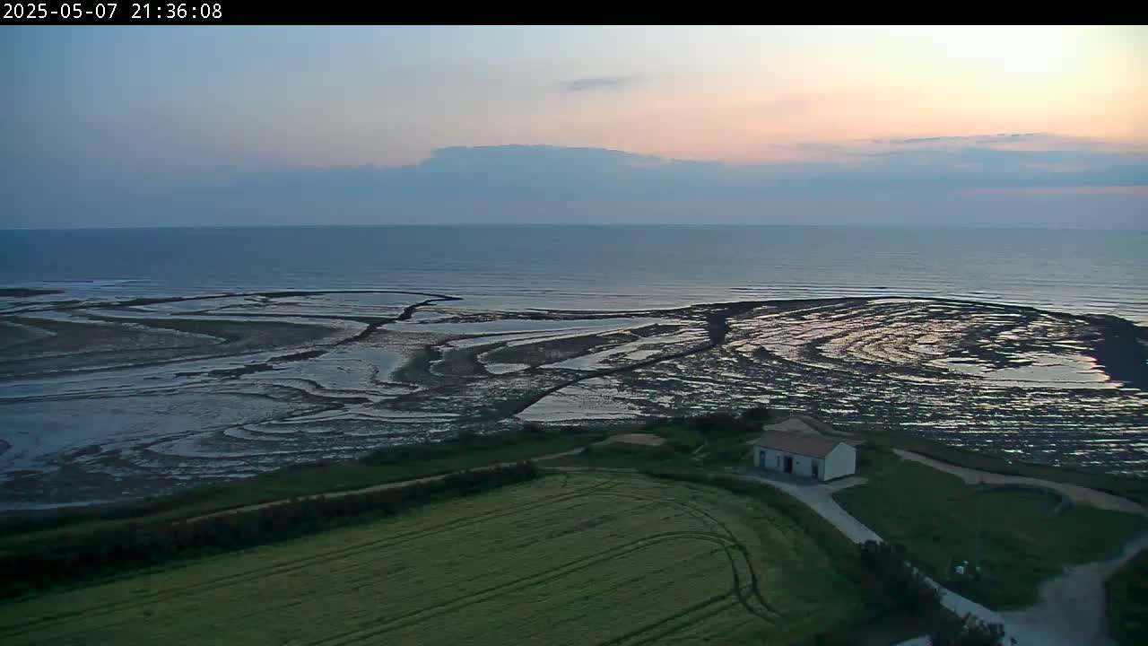 A small building sits on a grassy field overlooking an expanse of dark, wet tidal flats meeting a calm ocean under a twilight sky.