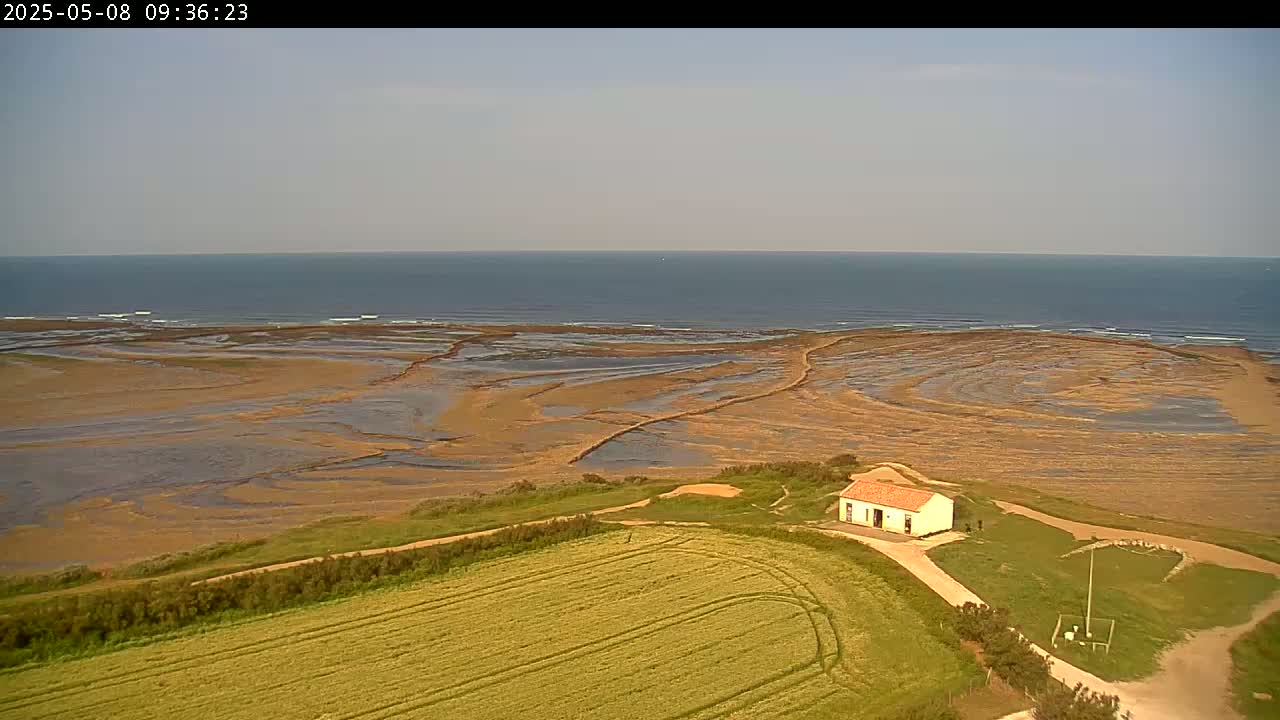 A small, white building sits on a grassy hill overlooking a vast, exposed tidal flat leading to a calm ocean under a mostly clear sky.