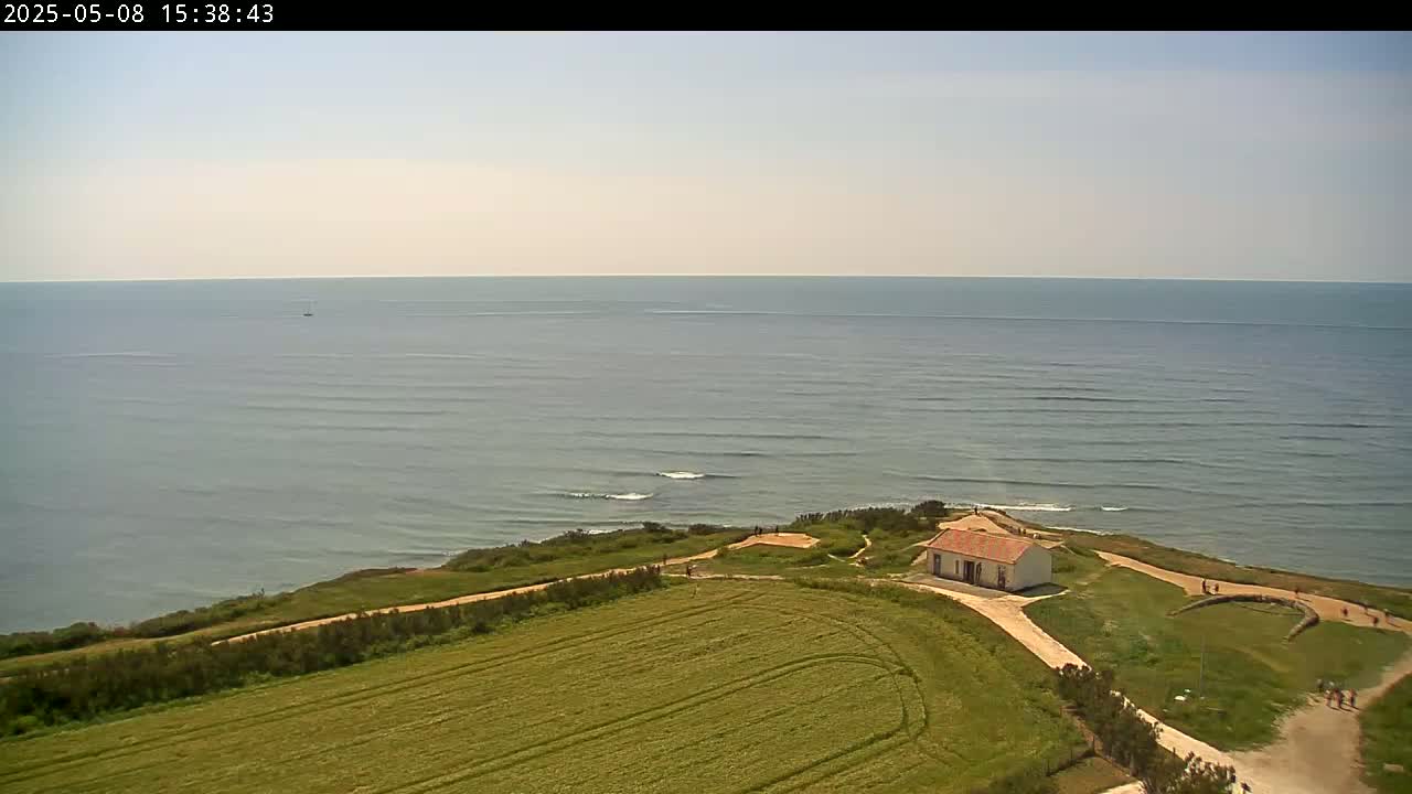 An aerial view shows a small, light-colored building on a grassy coastal bluff overlooking a calm ocean on a sunny day.