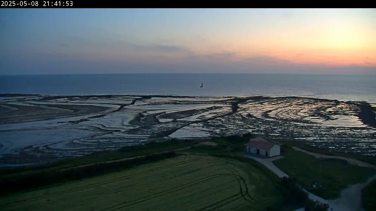 A small building sits on a grassy hill overlooking a tidal flat patterned with dark pools reflecting the sunset's orange and purple hues, with a calm sea in the background.