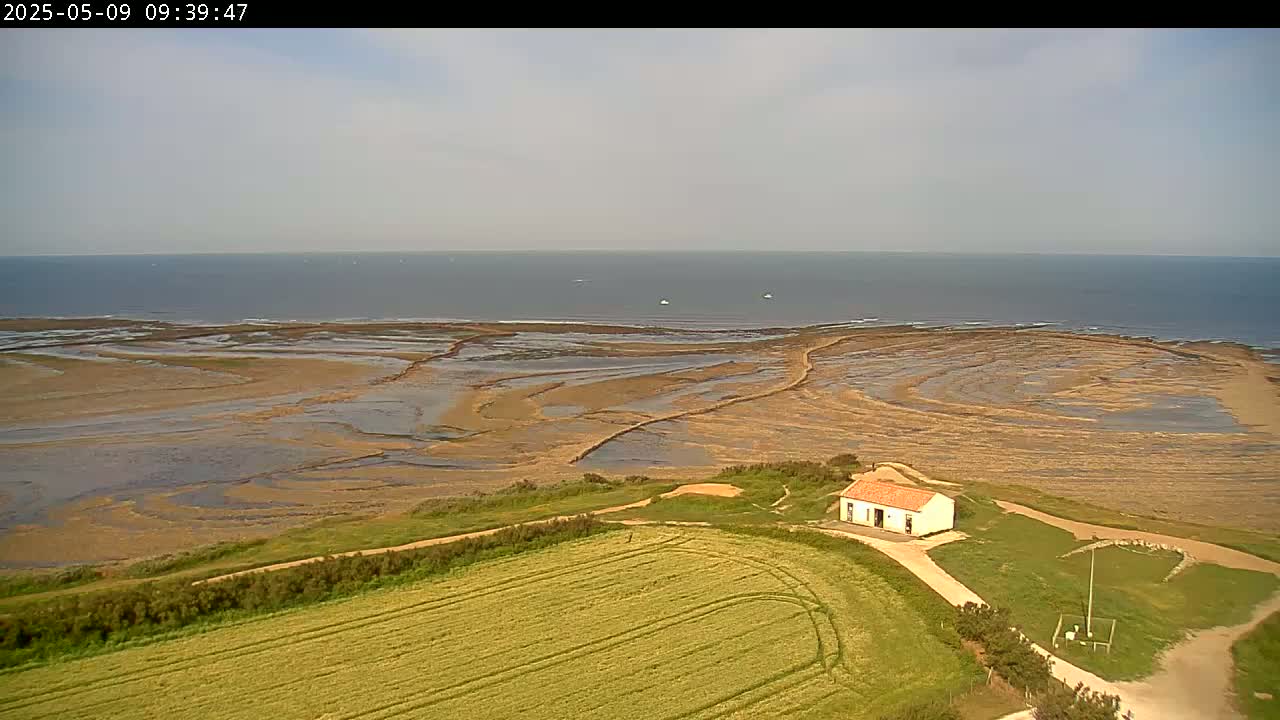 A small, white building sits on a grassy hill overlooking a wide expanse of tidal flats leading to a calm ocean under a mostly clear sky.