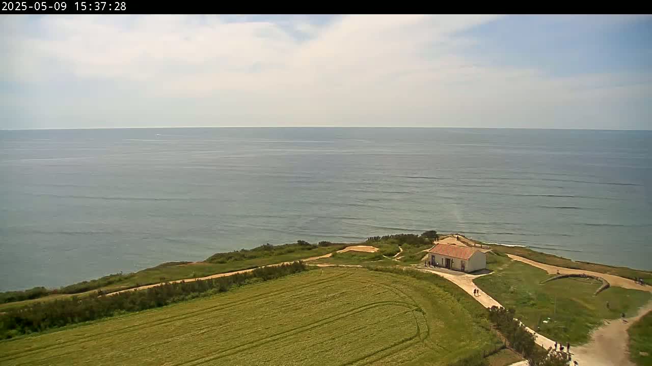 A small building sits on a grassy cliff overlooking a calm ocean under a mostly clear sky.