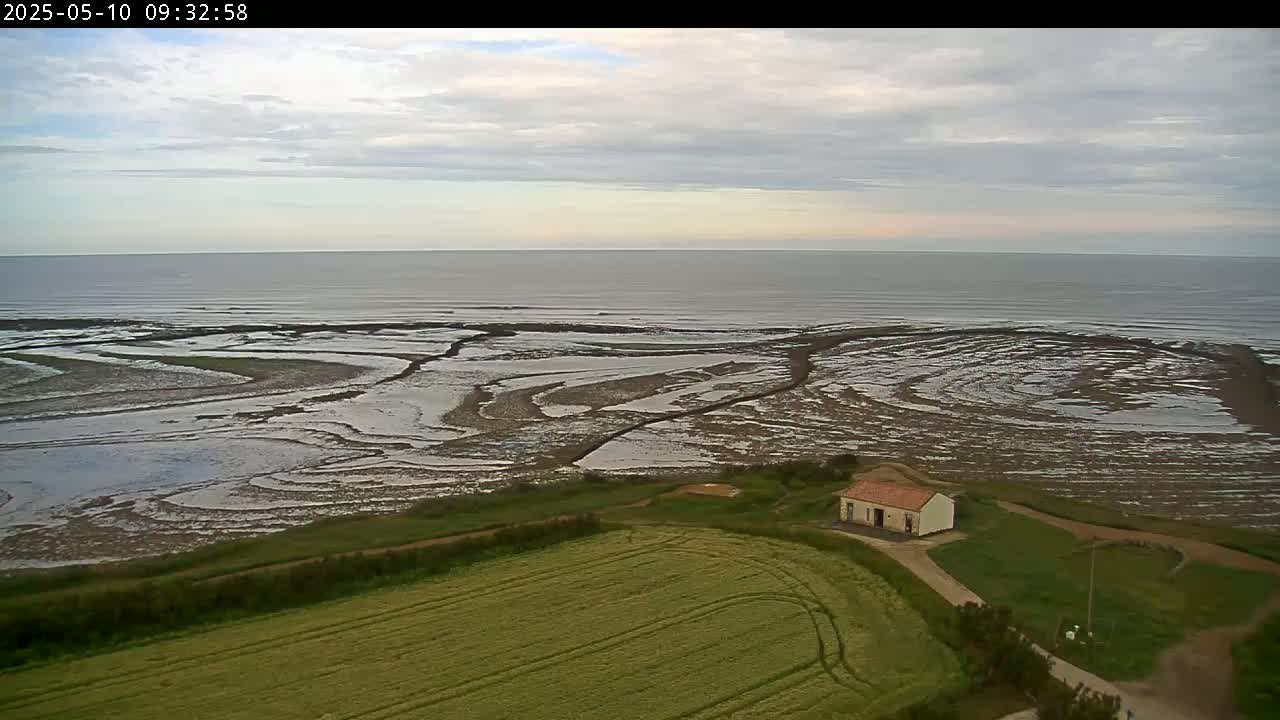 An aerial view shows a calm sea under a cloudy sky, with tidal flats and a small building situated near a field next to the water.