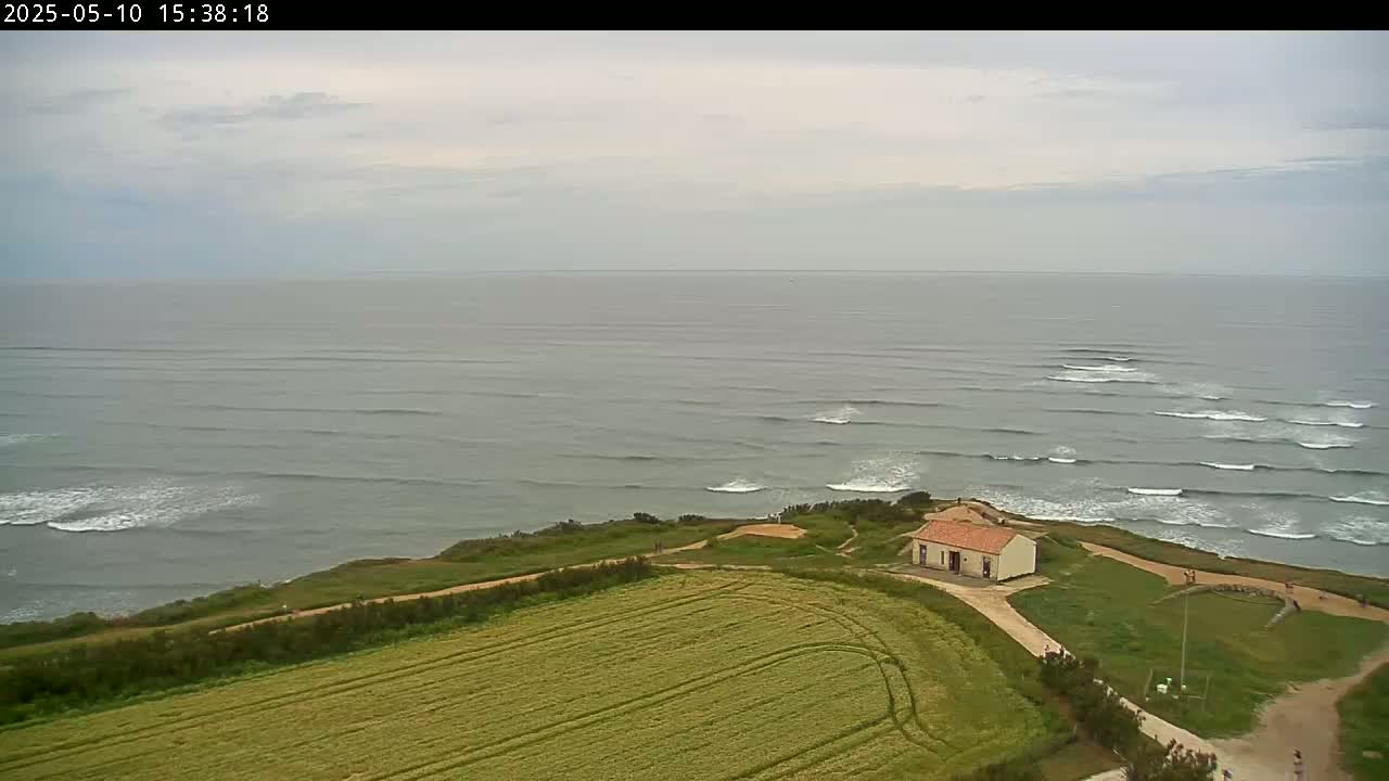 An aerial view shows a small building on a grassy cliff overlooking a gray, wavy ocean under an overcast sky.