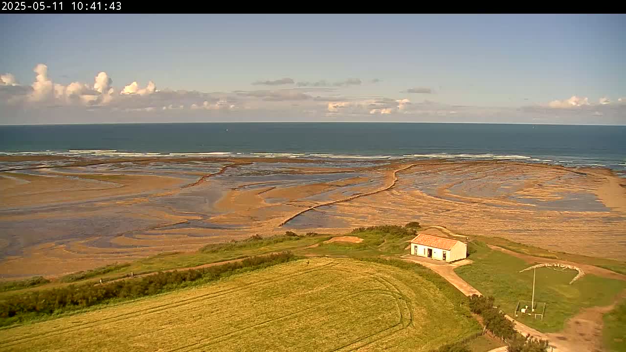A small building sits on a grassy hill overlooking a wide expanse of tan, wet sand and a calm ocean under a partly cloudy sky.