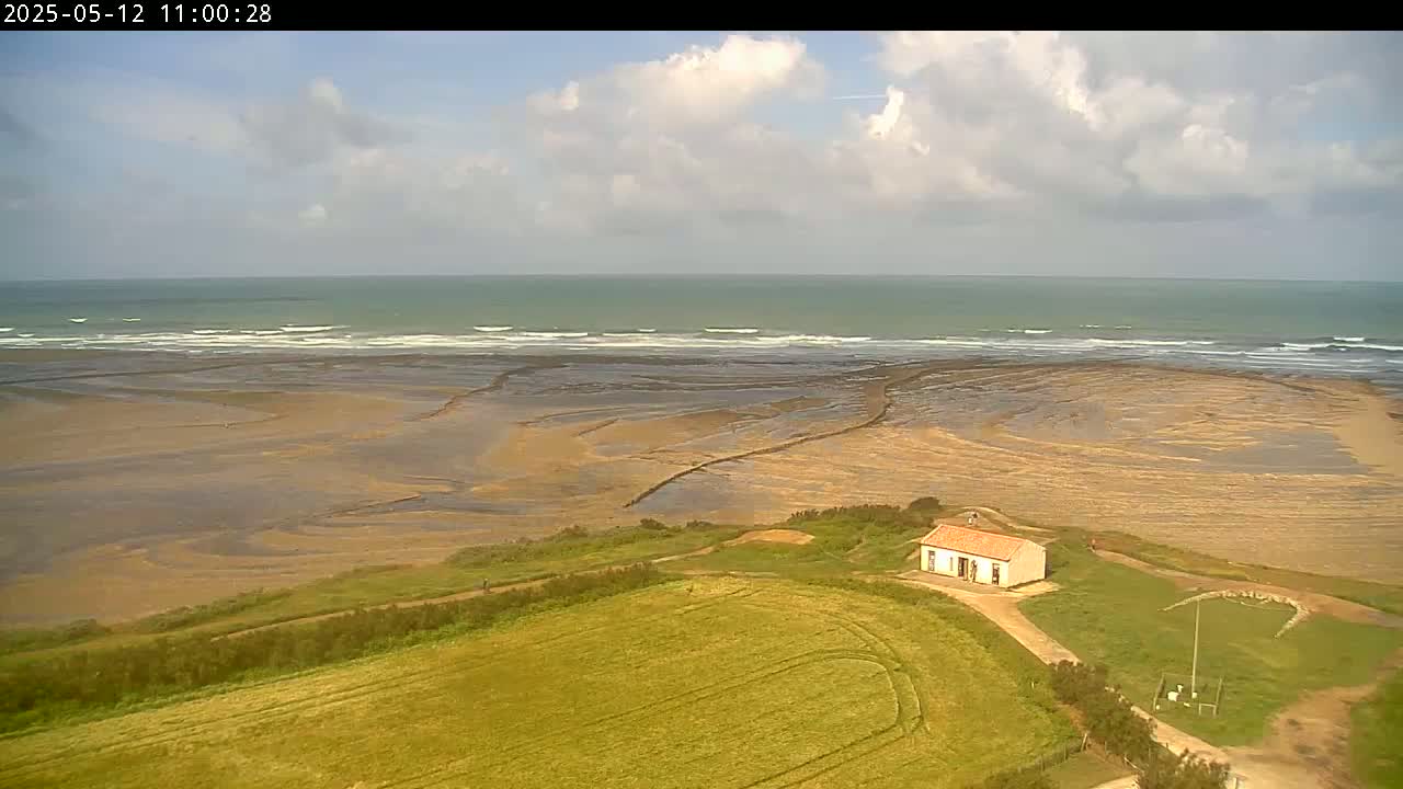 An aerial view shows a small building on a grassy hill overlooking a wide expanse of sandy beach and a partly cloudy sky above a gently-waving ocean.