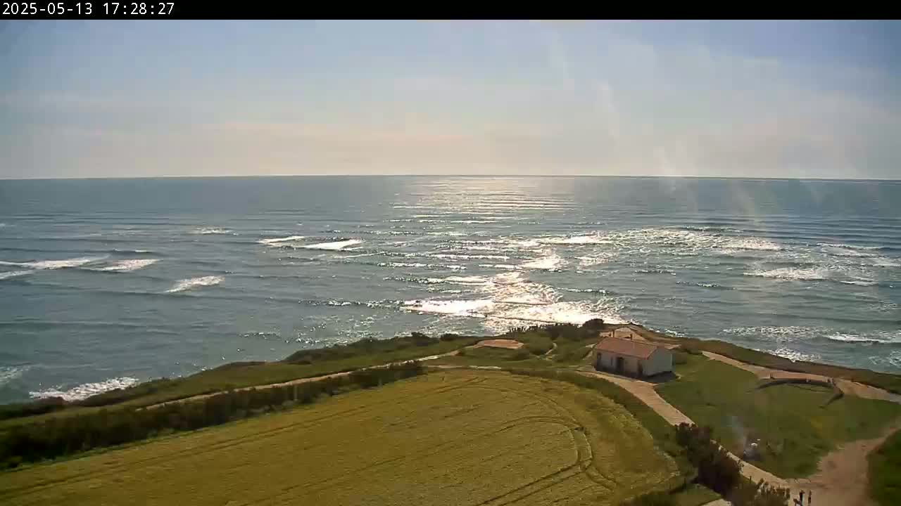A high-angle view shows a calm ocean with small waves under a mostly sunny sky, overlooking a grassy coastal area with a small building.
