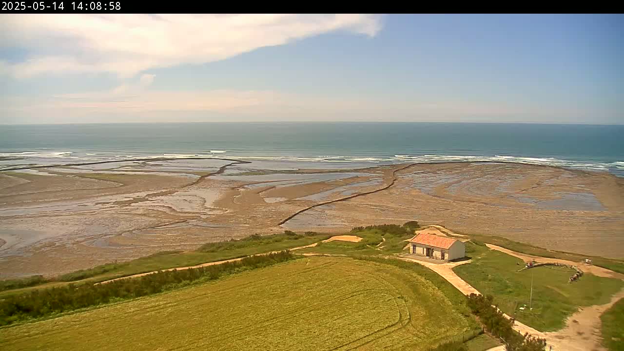 A small house sits on a grassy hill overlooking a tidal flat, with a calm ocean and light clouds visible in the background.