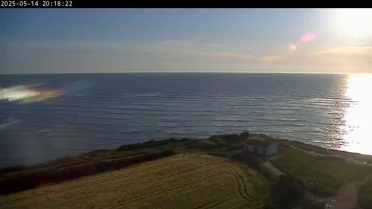 A high-angle view shows a calm ocean under a mostly clear sky at sunset, with a small house and a field visible on the shore.