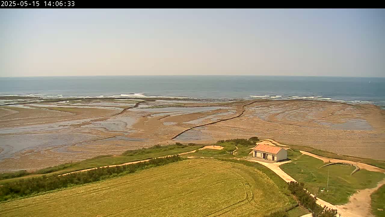 A small, white house with a red roof sits on a grassy hill overlooking a broad expanse of tidal flats and a calm ocean under a clear sky.