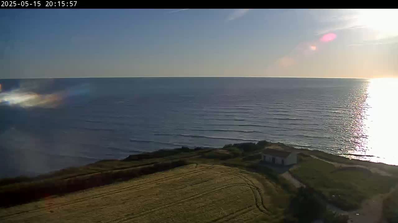 A high-angle view shows a calm ocean reflecting sunlight at sunset, with a small building nestled on a grassy hillside overlooking the water.