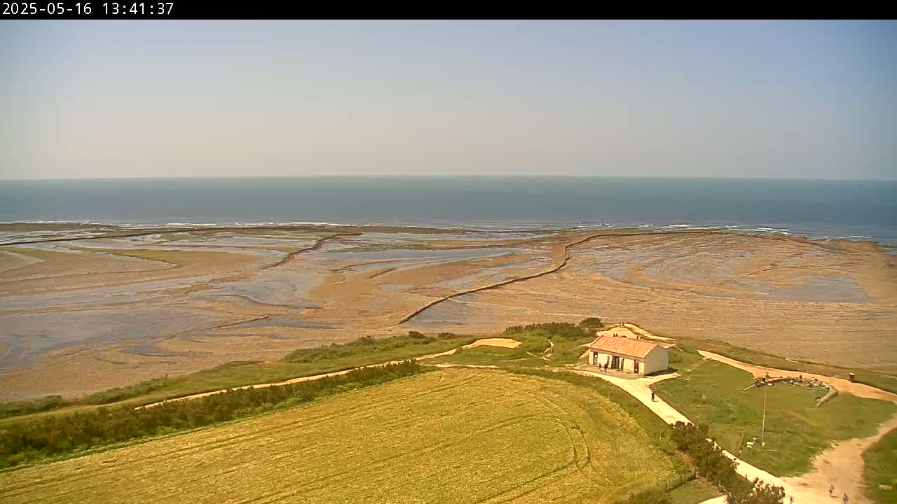 A small, white house sits on a grassy hill overlooking a tidal flat and calm ocean under a clear sky.