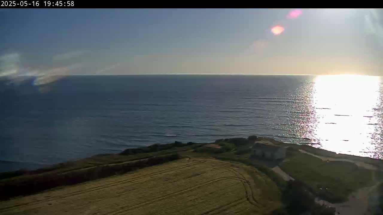 A high-angle view shows a calm ocean reflecting sunlight under a mostly clear sky, with a field and some structures visible on the land.