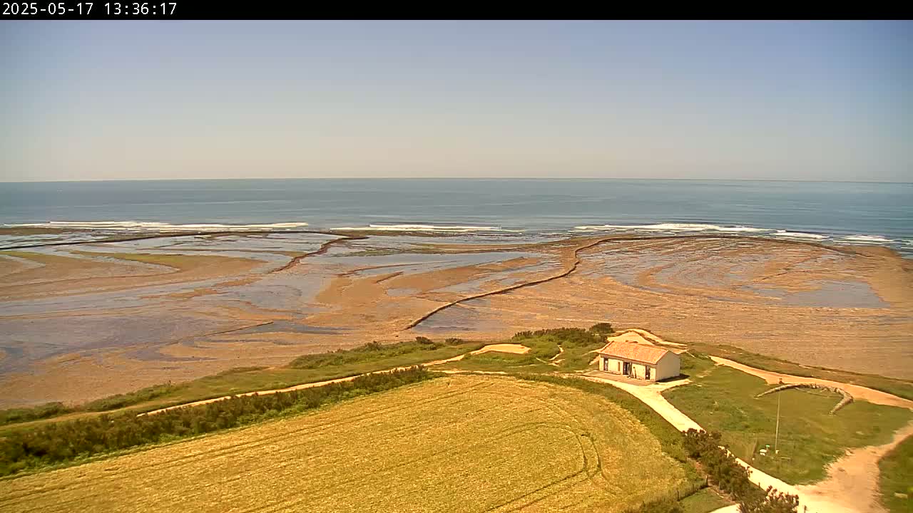 A small house sits on a grassy hill overlooking a sandy, tidal shoreline with gentle waves under a clear sky.