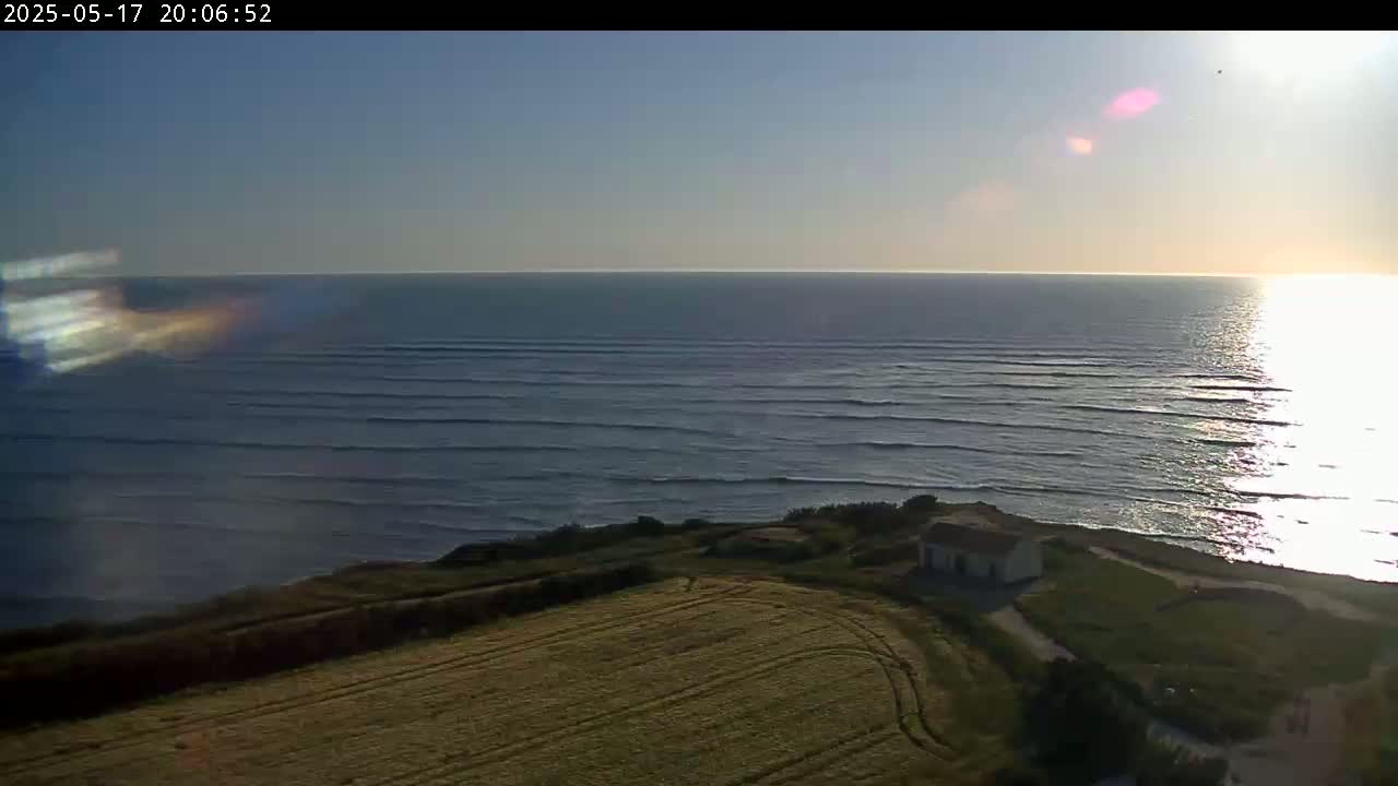 A small building sits atop a grassy cliff overlooking a calm ocean under a clear, sunny sky.