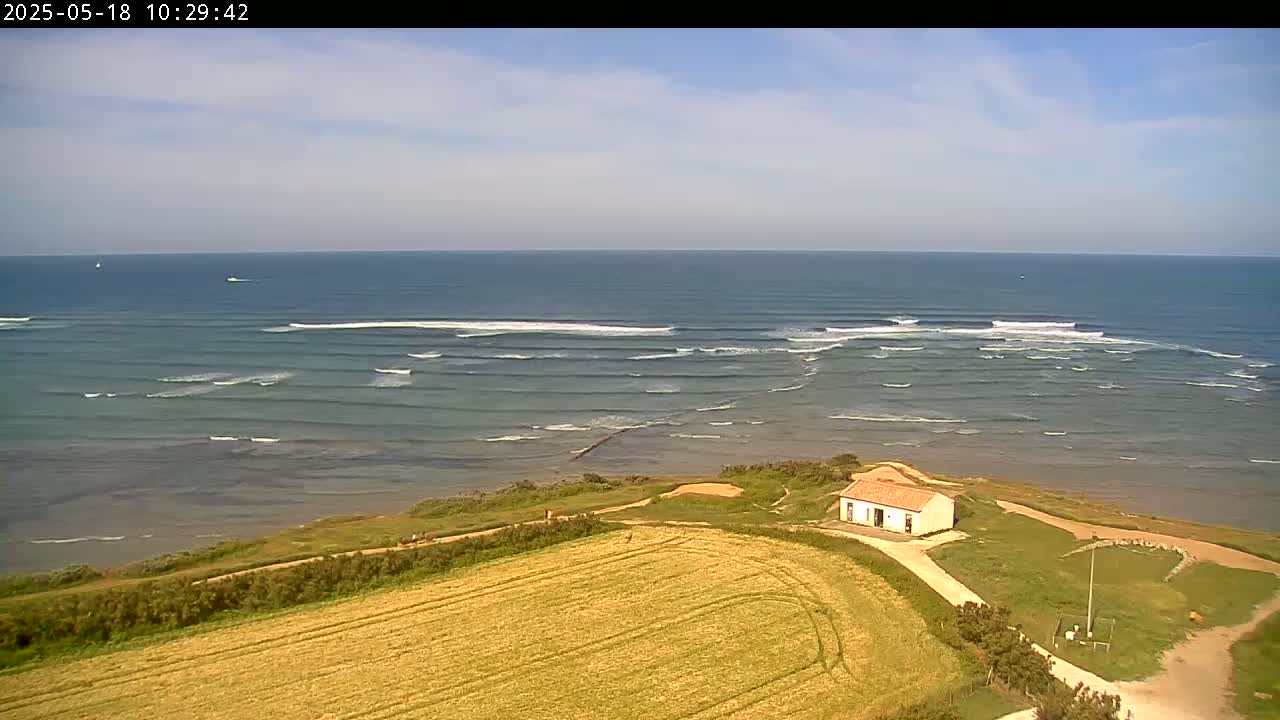 A high-angle view shows a small building near a gently sloping coastal field overlooking a calm ocean with small, rolling waves under a mostly clear sky.