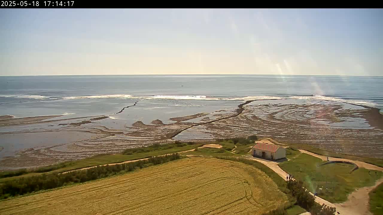 An aerial view shows a small building near a gently sloping field overlooking a rocky, partially submerged shoreline and calm ocean under a sunny sky.