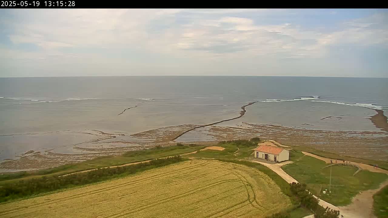A small, single-story building sits on a grassy hill overlooking a calm ocean with some small waves breaking near a rocky outcropping under a partly cloudy sky.