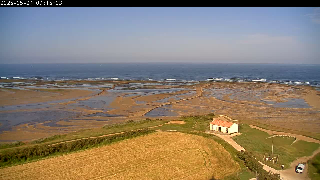 A small house sits near a coastal field overlooking a calm ocean and exposed tidal flats under a clear sky.