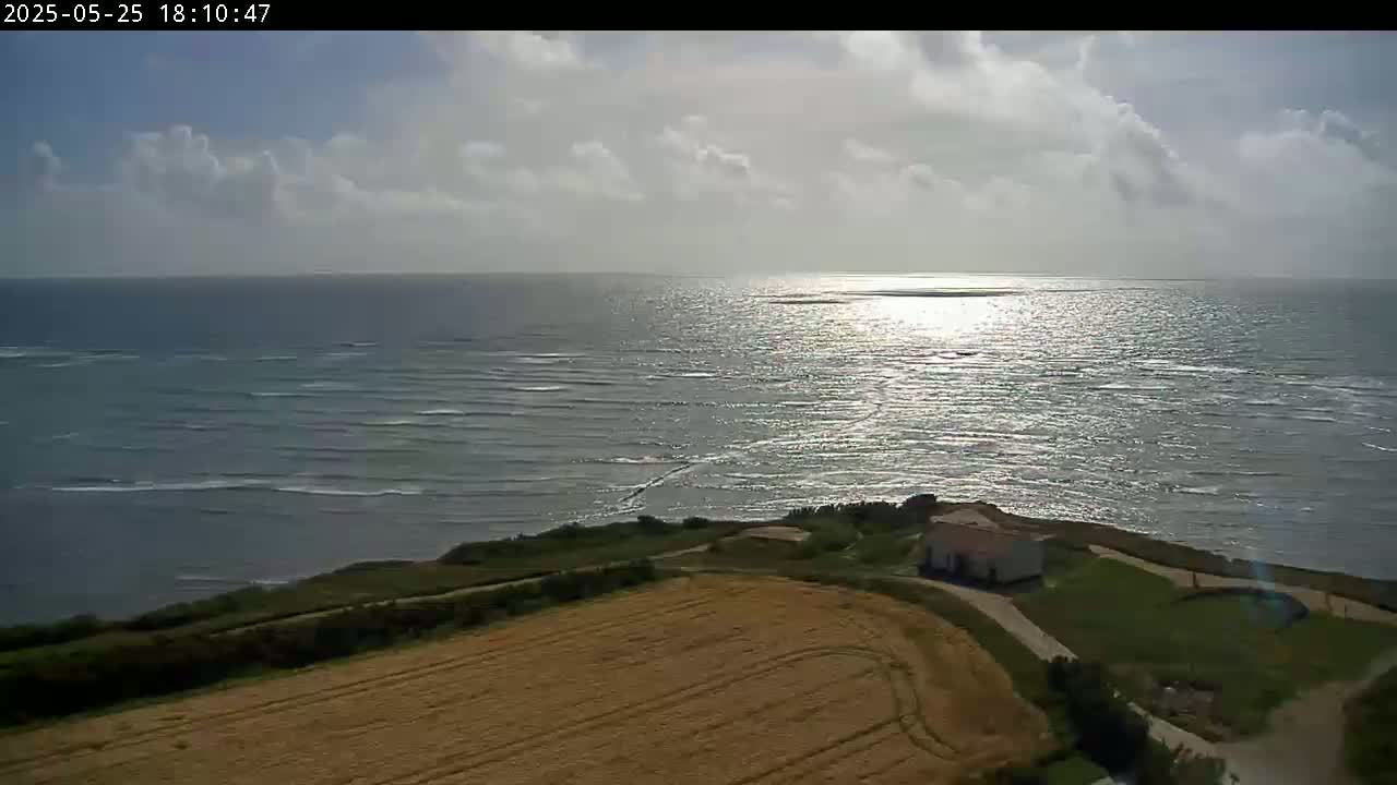 A partly cloudy day shows a view of a calm sea reflecting sunlight, with a small building nestled on a grassy cliff overlooking a golden field.