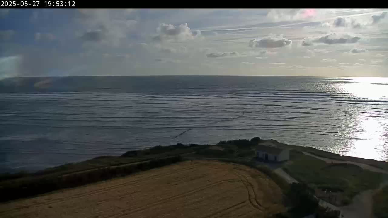A partly cloudy sunset over a wavy ocean, viewed from a high vantage point overlooking a field and a small building near a cliff.