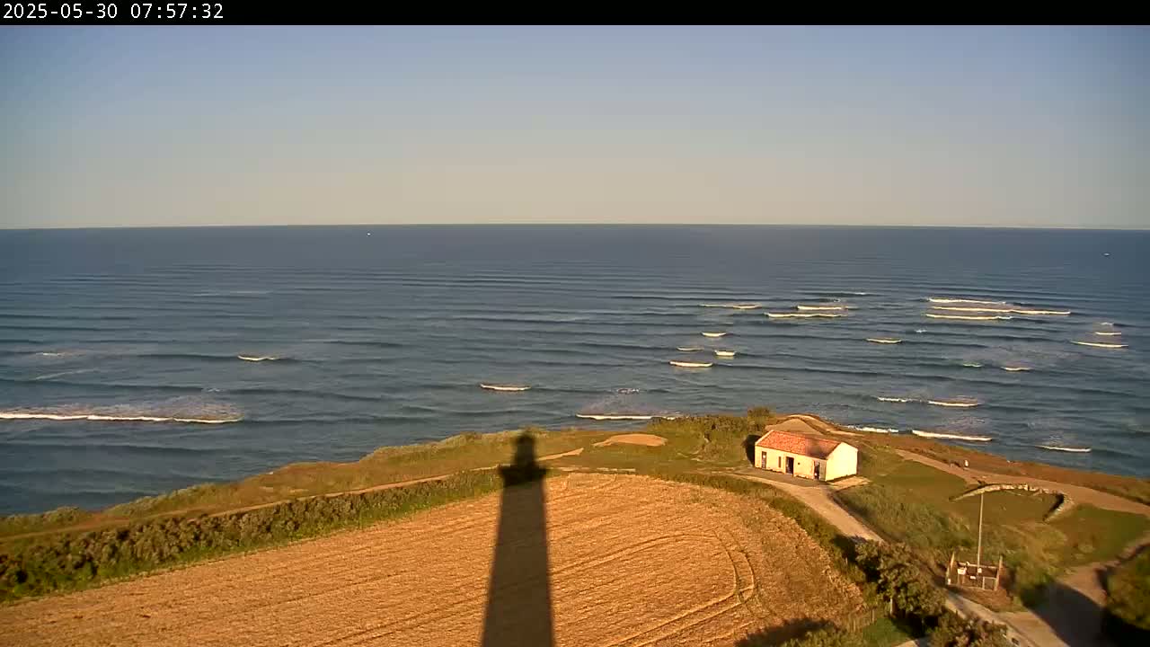 A long shadow from a tall structure stretches across a golden field overlooking a calm ocean with small waves breaking near the shore, under a clear sky.