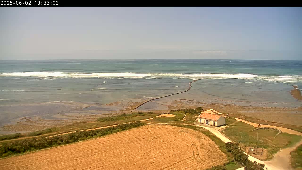 A small house sits on a tan field overlooking a calm ocean with small waves breaking near the shore under a clear sky.