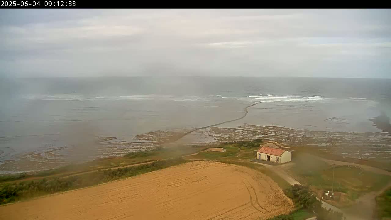 A small building sits near a brown field overlooking a hazy ocean with small waves under a cloudy sky.