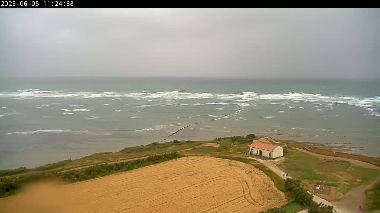 A small, light-colored building sits on a grassy coastal area overlooking a choppy sea under an overcast sky.