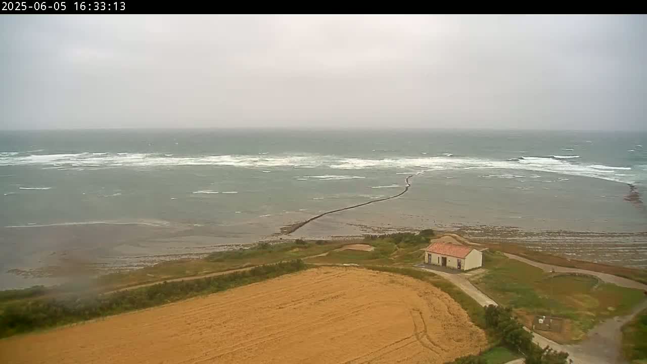 A high-angle view shows a small building near a harvested field next to a choppy sea under an overcast sky.