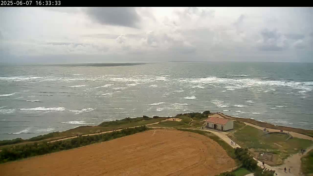 A coastal scene under a partly cloudy sky shows a choppy gray sea meeting a grassy shoreline with a small white building and a brown field.