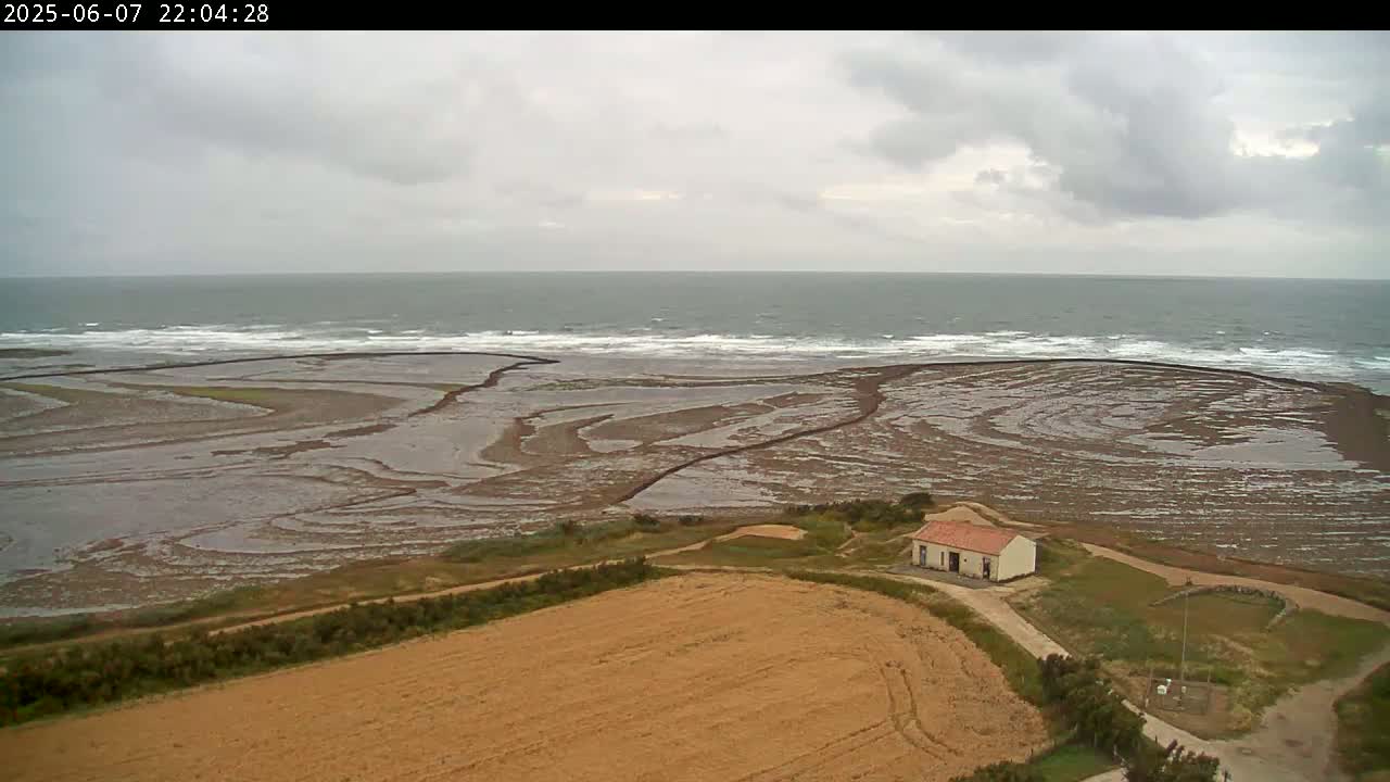 An aerial view shows a small building next to a brown field overlooking a tidal flat and a choppy gray sea under an overcast sky.