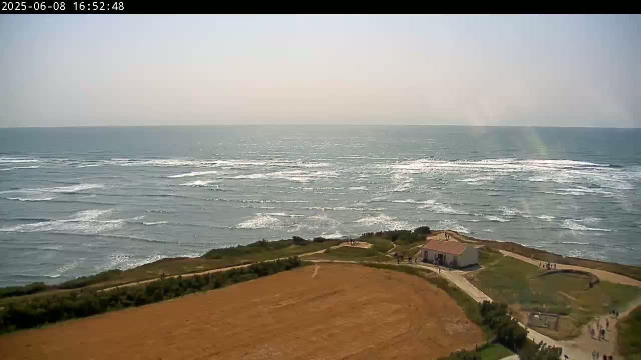 A mostly sunny day shows a view of a rocky coastline with a small building and a brown field overlooking a wavy ocean.