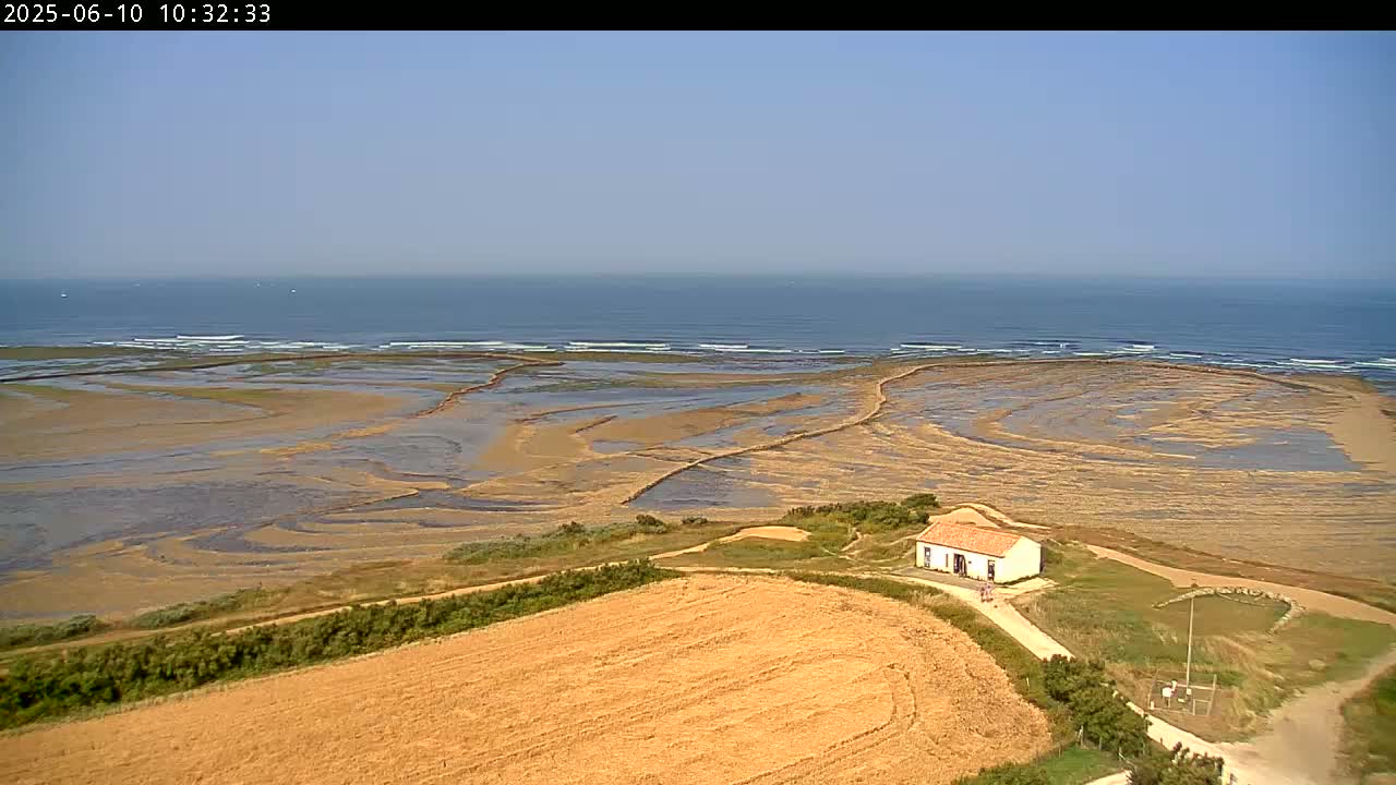 A small white house sits on a grassy hill overlooking a wide expanse of tidal flats that lead to a calm ocean under a clear sky.