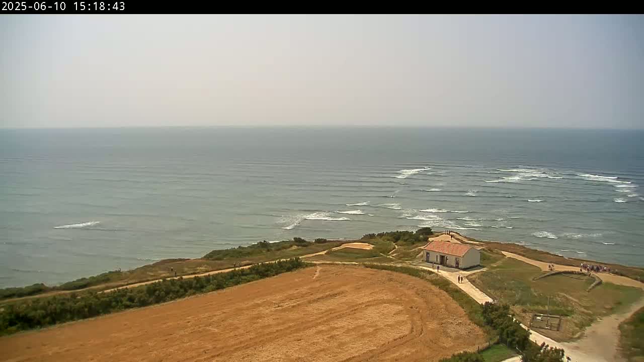 A small, light-colored building sits atop a grassy cliff overlooking a calm ocean with small waves under a hazy sky.