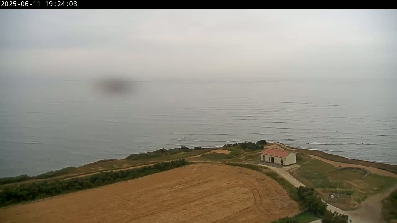 A hazy coastal scene shows a small building nestled on a grassy cliff overlooking a calm sea and a tan field.