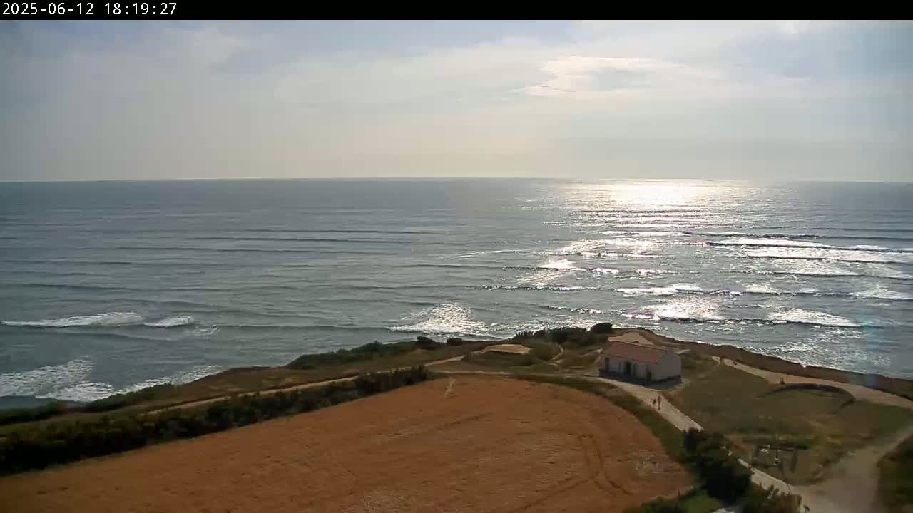 A partly sunny day shows a view of a calm ocean with small waves, a small building near a grassy cliff overlooking a tan field.