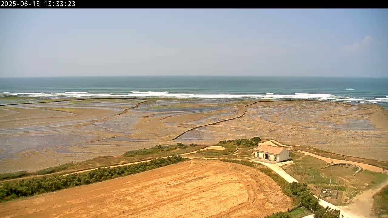 A small house sits on a dry, tan field overlooking a tidal flat and a light-blue ocean with small waves under a clear sky.