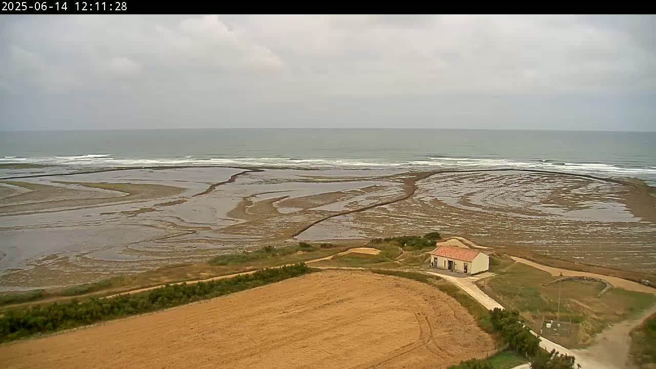 A small, light-colored building sits near a vast expanse of tidal flats and a calm, gray ocean under an overcast sky.