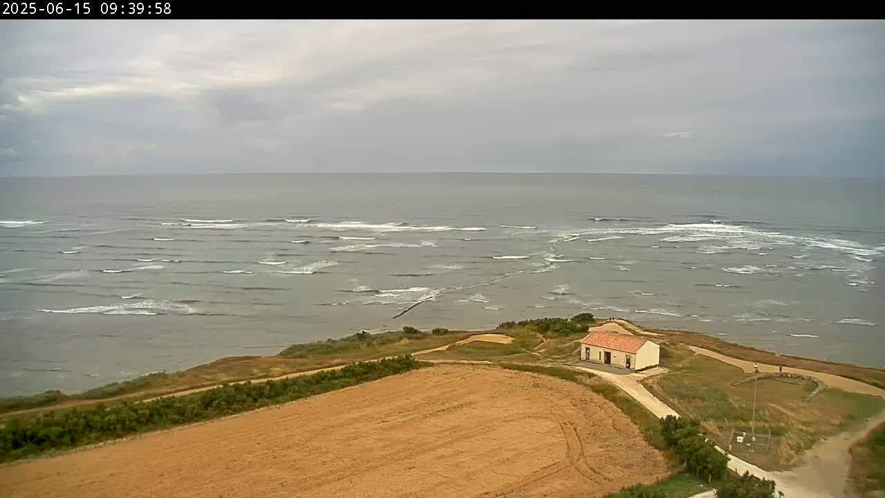 An aerial view shows a small building on a grassy cliff overlooking a calm sea with small waves under an overcast sky.