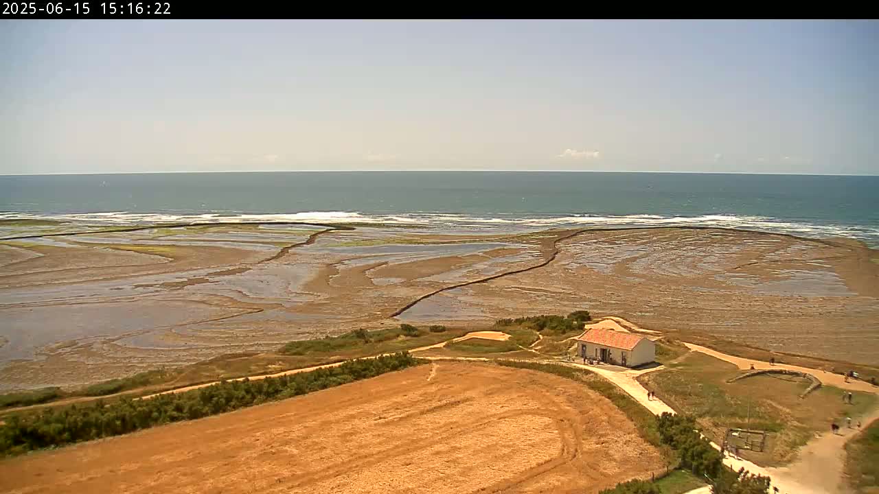 A high-angle view shows a calm ocean under a mostly clear sky, a tidal flat with exposed seabed patterns, and a small building situated on a grassy area next to a dry, tan field.