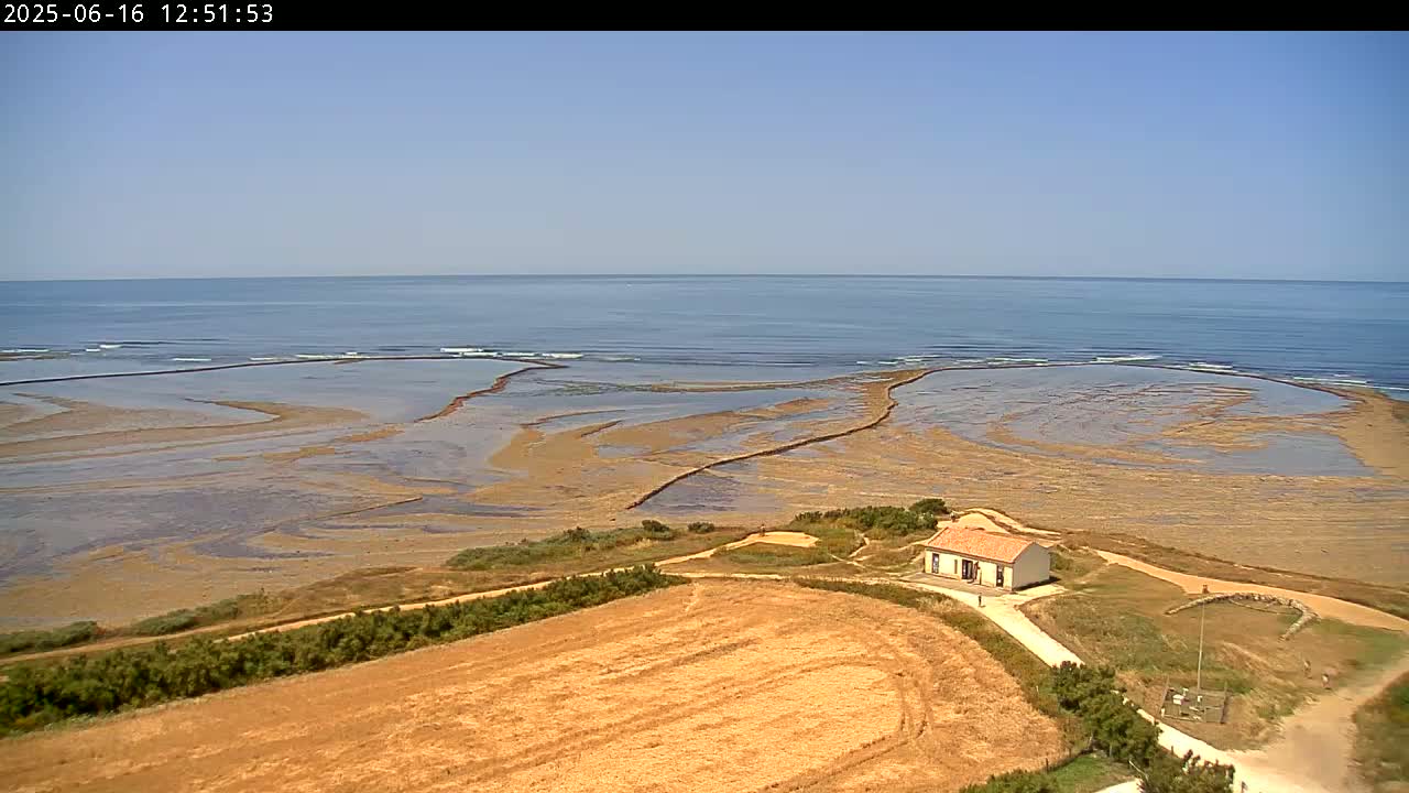 An aerial view shows a small house situated on a coastal path overlooking a calm ocean and expansive tidal flats under a clear sky.
